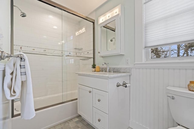 full bathroom with a wainscoted wall, combined bath / shower with glass door, toilet, and vanity