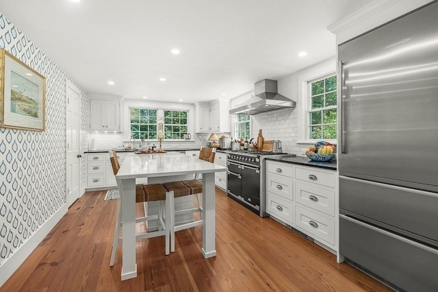 kitchen featuring wall chimney range hood, premium appliances, wallpapered walls, a breakfast bar, and white cabinetry
