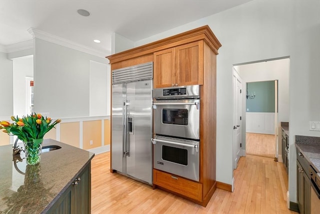 kitchen featuring brown cabinetry, crown molding, light wood-style floors, dark stone counters, and appliances with stainless steel finishes