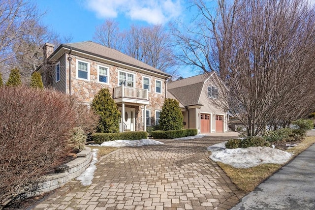 view of front facade with a balcony, a garage, stone siding, decorative driveway, and a chimney