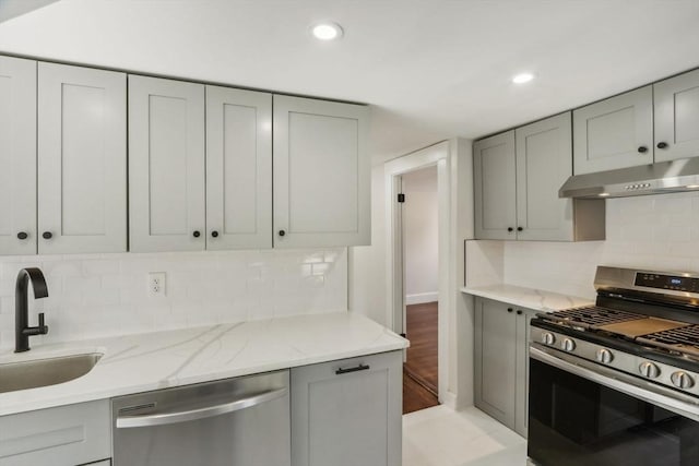 kitchen featuring light stone countertops, under cabinet range hood, gray cabinets, stainless steel appliances, and a sink