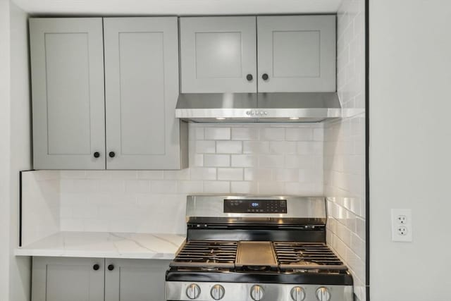 kitchen with stainless steel gas range oven, gray cabinetry, under cabinet range hood, decorative backsplash, and light stone countertops