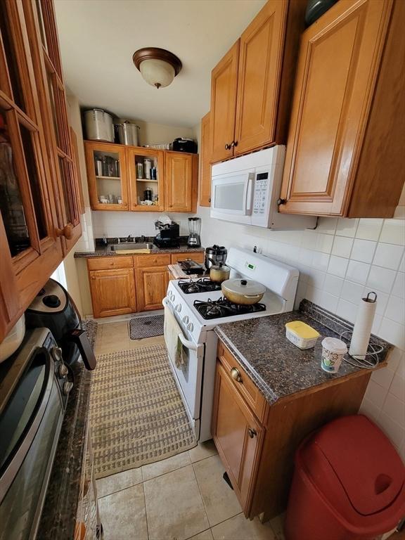 kitchen with white appliances, brown cabinetry, light tile patterned flooring, a sink, and glass insert cabinets