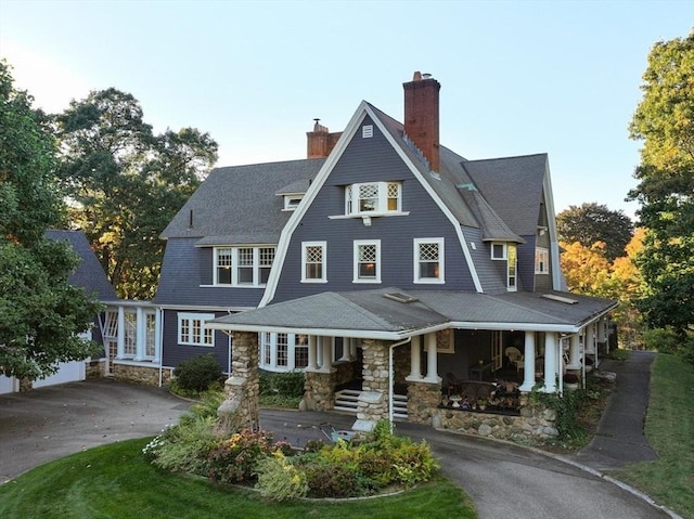 view of front facade featuring a porch, driveway, a chimney, a gambrel roof, and stone siding