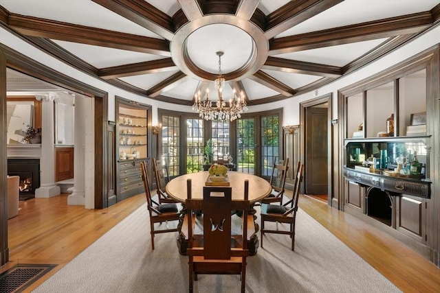 dining area featuring coffered ceiling, a chandelier, ornamental molding, a warm lit fireplace, and light wood finished floors