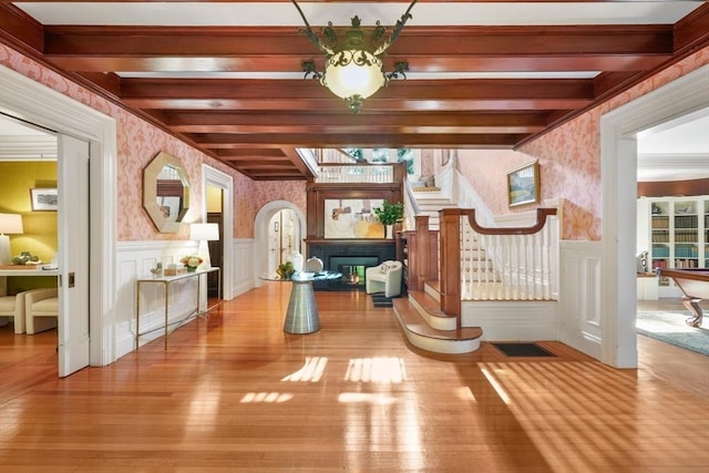 interior space with wallpapered walls, a wainscoted wall, beamed ceiling, wood finished floors, and a glass covered fireplace