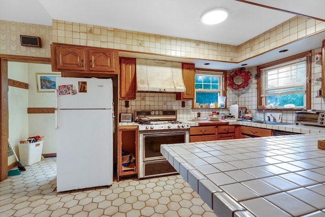 kitchen with decorative backsplash, range with gas cooktop, wall chimney range hood, white refrigerator, and tile counters