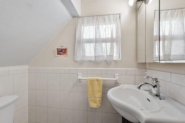 bathroom featuring toilet, a wainscoted wall, lofted ceiling, a sink, and tile walls