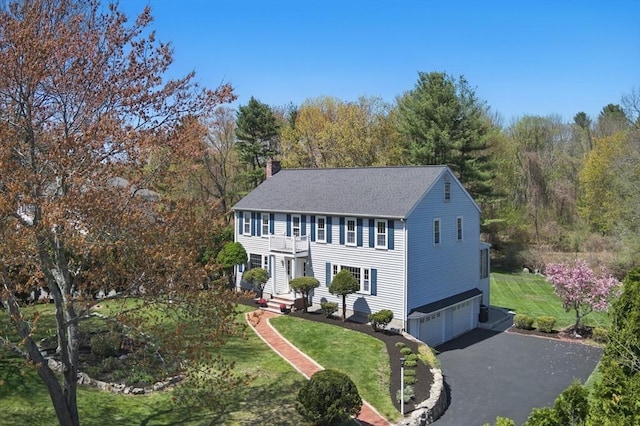 colonial home with a chimney, a garage, a front lawn, roof with shingles, and driveway