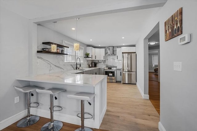 kitchen with light wood finished floors, appliances with stainless steel finishes, a sink, a peninsula, and wall chimney exhaust hood