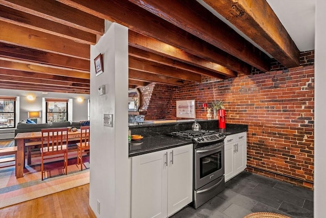 kitchen featuring stainless steel gas range, white cabinets, brick wall, dark stone counters, and beamed ceiling