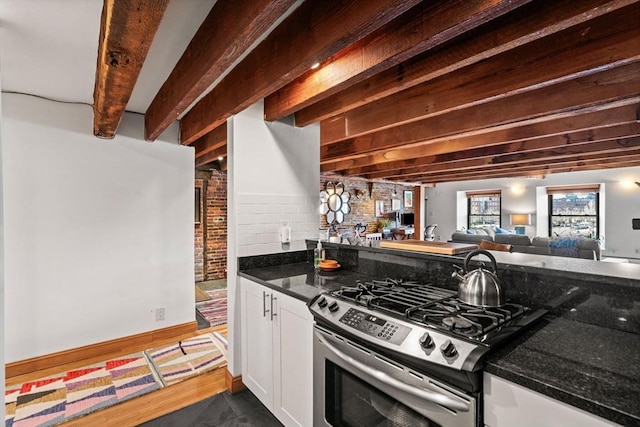 kitchen featuring gas range, beamed ceiling, dark stone counters, and white cabinets