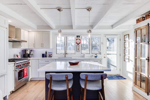 kitchen with pendant lighting, beamed ceiling, white cabinets, stainless steel range, and wall chimney exhaust hood