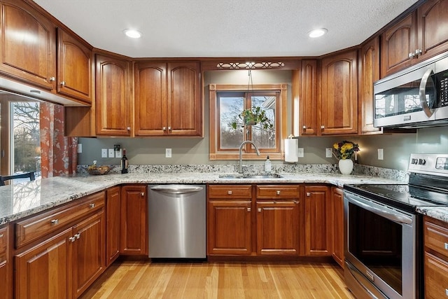 kitchen with appliances with stainless steel finishes, a wealth of natural light, sink, light stone countertops, and light wood-type flooring