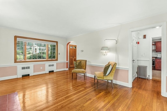 sitting room with radiator heating unit, radiator, arched walkways, light wood-type flooring, and crown molding