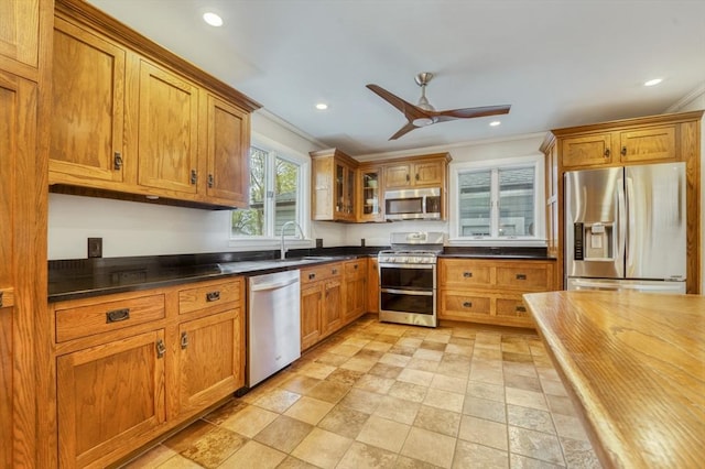 kitchen with brown cabinets, dark countertops, appliances with stainless steel finishes, a ceiling fan, and ornamental molding