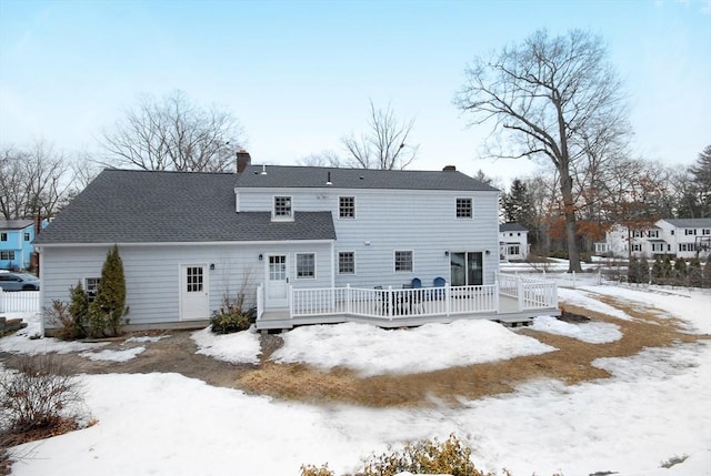 snow covered house featuring a wooden deck, roof with shingles, and a chimney