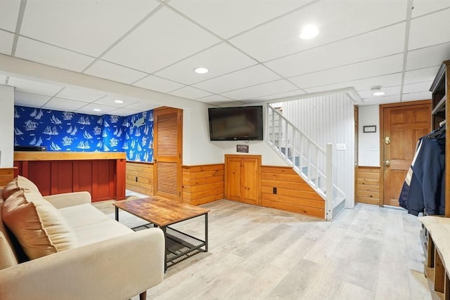living room featuring wainscoting, wallpapered walls, stairway, and a drop ceiling