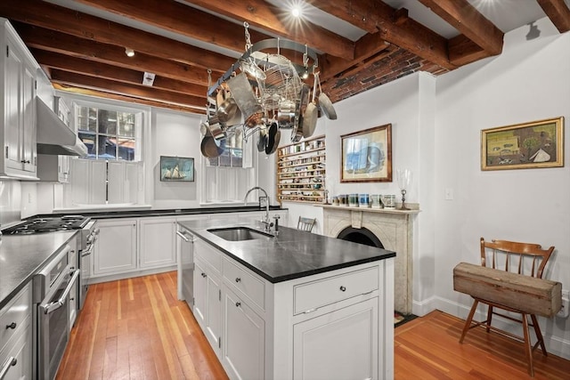 kitchen with a sink, appliances with stainless steel finishes, light wood-style flooring, dark countertops, and beamed ceiling