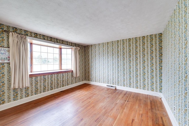 empty room featuring wood-type flooring, baseboards, wallpapered walls, and a textured ceiling