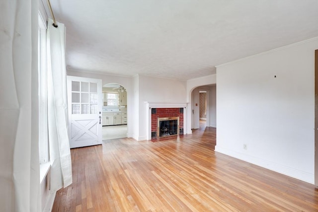 unfurnished living room featuring arched walkways, light wood-type flooring, ornamental molding, a brick fireplace, and baseboards