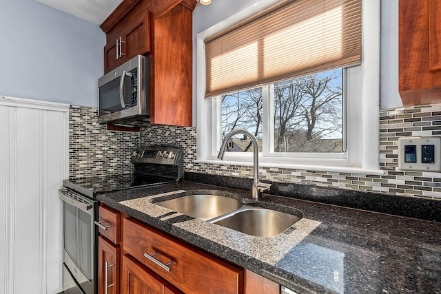 kitchen featuring brown cabinets, backsplash, appliances with stainless steel finishes, a sink, and dark stone counters