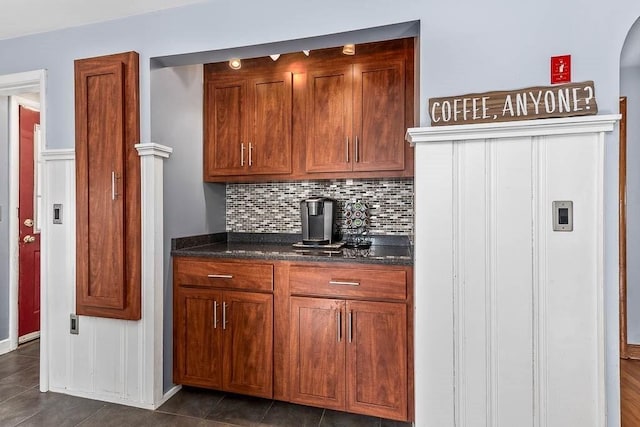 kitchen with dark tile patterned flooring, brown cabinets, backsplash, and dark stone countertops