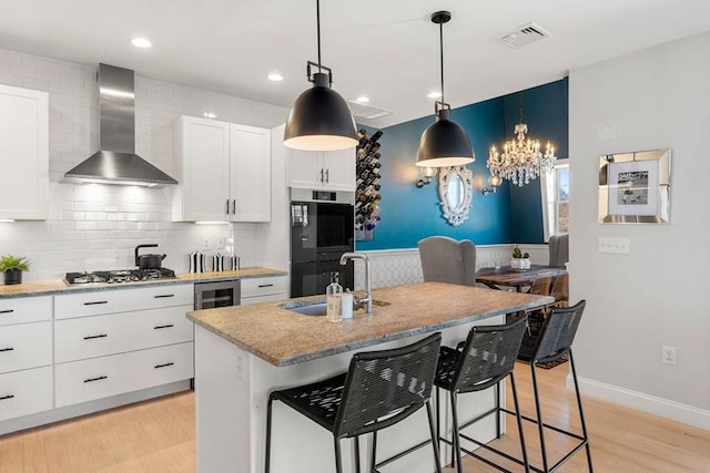 kitchen with beverage cooler, a sink, double oven, wall chimney exhaust hood, and light wood-type flooring