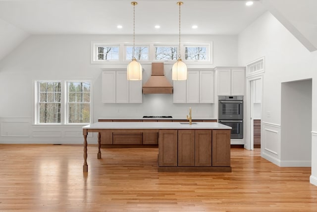 kitchen with white cabinetry, wall chimney exhaust hood, light hardwood / wood-style flooring, a center island with sink, and double oven