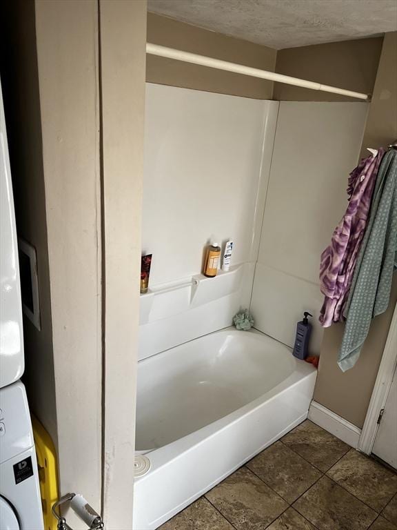 bathroom with a textured ceiling, a tub, baseboards, and stacked washer and dryer