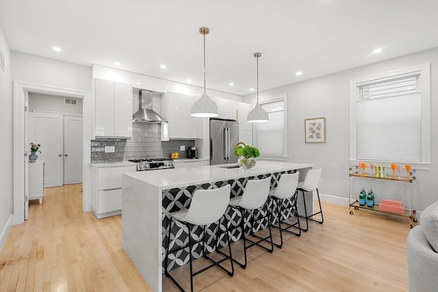 kitchen with hanging light fixtures, wall chimney exhaust hood, an island with sink, high end fridge, and white cabinetry