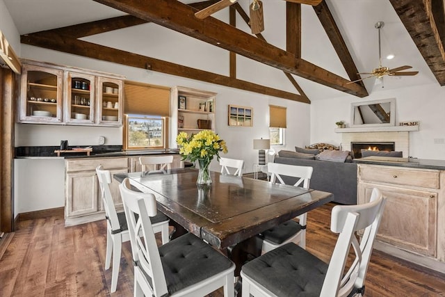 dining space featuring ceiling fan, high vaulted ceiling, a lit fireplace, dark wood-style floors, and beamed ceiling