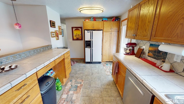 kitchen featuring a textured ceiling, white refrigerator with ice dispenser, hanging light fixtures, tile counters, and dishwasher