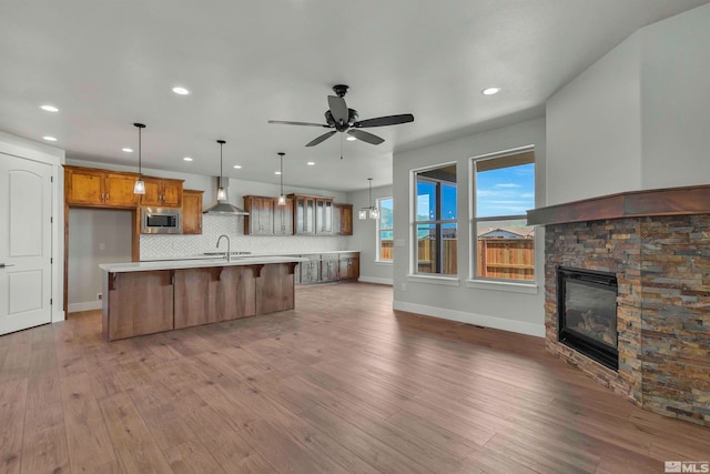 kitchen with stainless steel microwave, ceiling fan, wood-type flooring, an island with sink, and wall chimney range hood