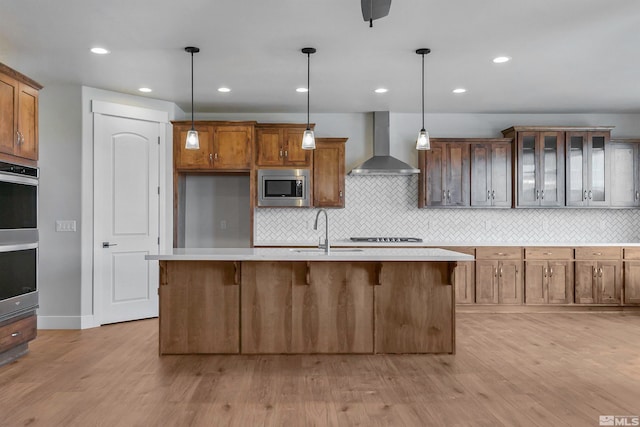 kitchen featuring appliances with stainless steel finishes, wall chimney range hood, an island with sink, backsplash, and light hardwood / wood-style flooring