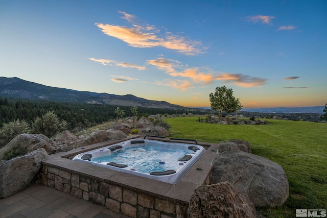 pool at dusk with a mountain view, a yard, and an outdoor hot tub