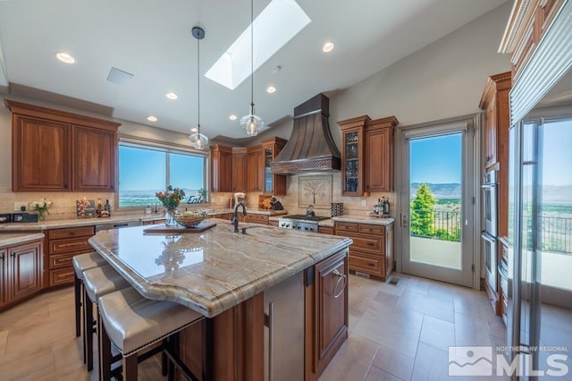 kitchen featuring tasteful backsplash, a skylight, light tile floors, an island with sink, and premium range hood