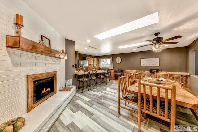 dining room featuring ceiling fan, a brick fireplace, light hardwood / wood-style floors, and a skylight