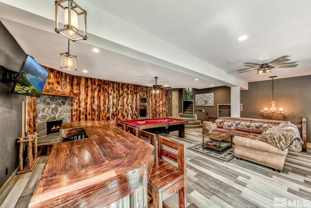 rec room with pool table, ceiling fan with notable chandelier, light wood-type flooring, wood walls, and a stone fireplace