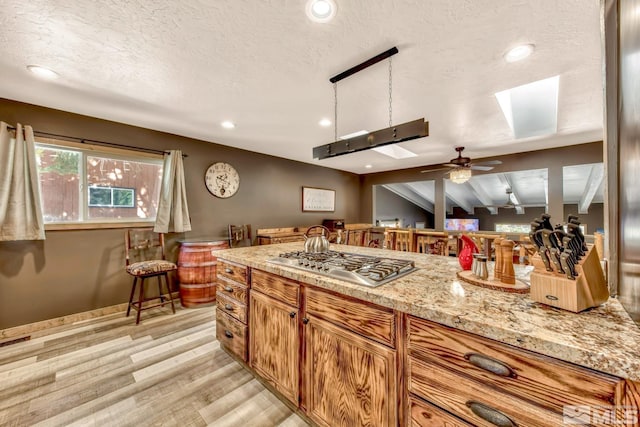 kitchen with light stone countertops, stainless steel gas cooktop, ceiling fan, light wood-type flooring, and lofted ceiling with skylight