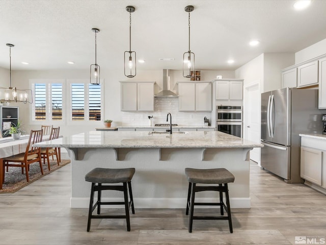 kitchen featuring decorative light fixtures, appliances with stainless steel finishes, a center island with sink, light wood-type flooring, and wall chimney range hood