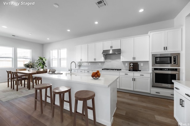 kitchen with an island with sink, dark hardwood / wood-style flooring, backsplash, stainless steel appliances, and white cabinetry