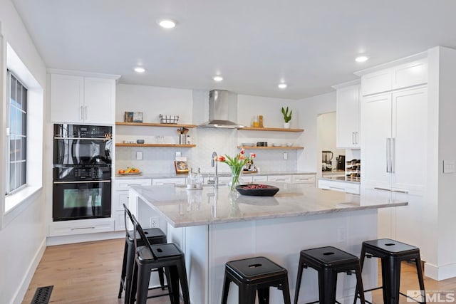 kitchen featuring light stone counters, white cabinets, light hardwood / wood-style flooring, double oven, and wall chimney exhaust hood