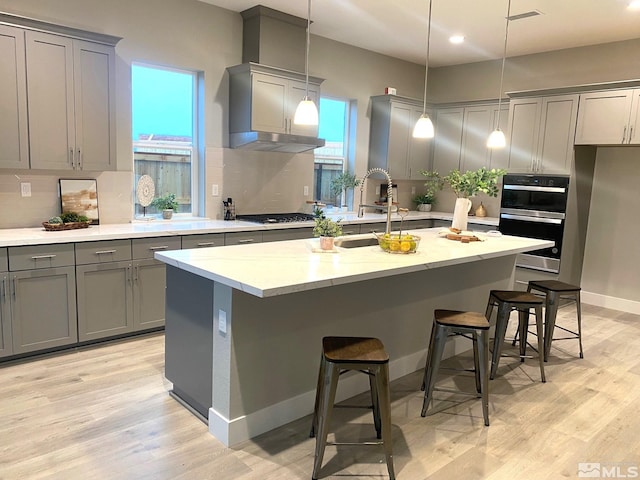 kitchen featuring dobule oven black, gray cabinetry, light wood-style floors, backsplash, and an island with sink