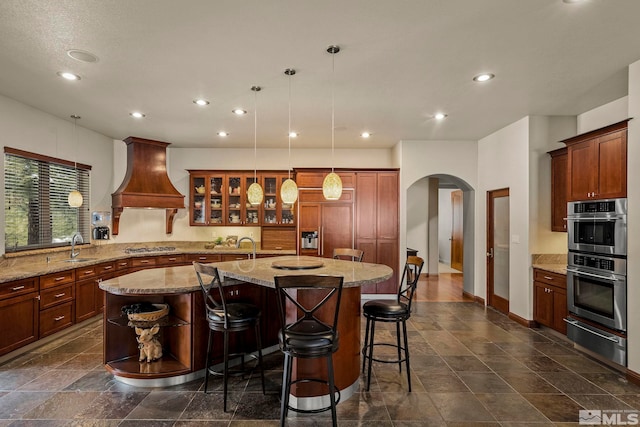 kitchen featuring custom range hood, appliances with stainless steel finishes, light stone counters, decorative light fixtures, and a kitchen island with sink