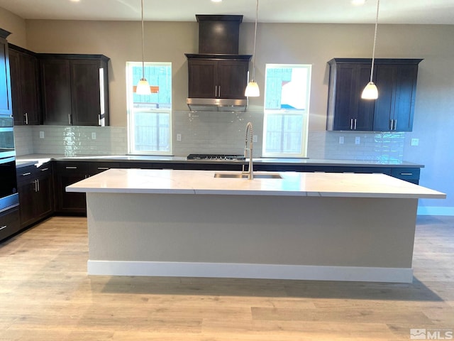 kitchen featuring light wood finished floors, wall chimney exhaust hood, oven, a kitchen island with sink, and a sink