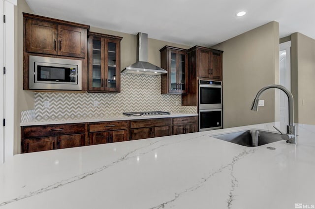 kitchen with backsplash, appliances with stainless steel finishes, wall chimney range hood, sink, and light stone counters