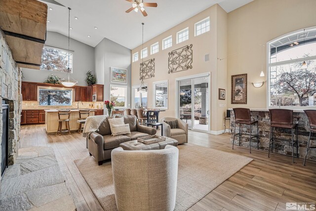 living room featuring high vaulted ceiling, ceiling fan, light hardwood / wood-style floors, and a stone fireplace