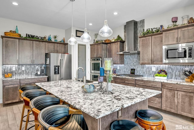 kitchen with appliances with stainless steel finishes, wall chimney range hood, tasteful backsplash, an island with sink, and a kitchen breakfast bar