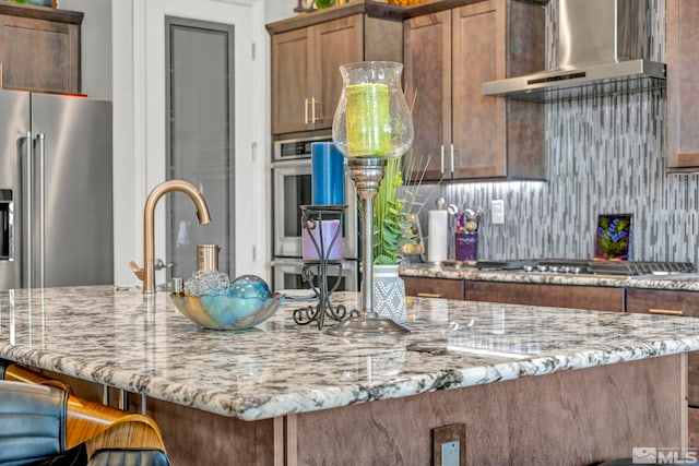 kitchen with appliances with stainless steel finishes, wall chimney exhaust hood, tasteful backsplash, an island with sink, and light stone countertops